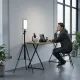 Mini lampe vidéo LED 40W portable avec batterie intégrée et charge PD pour studio et création de contenu Mini lampe vidéo LED 40W portable avec batterie intégrée et charge PD posée sur un trépied à côté d'un jeune homme assis à un bureau équipé d'un micro, d'un appareil photo et d'un ordinateur portable